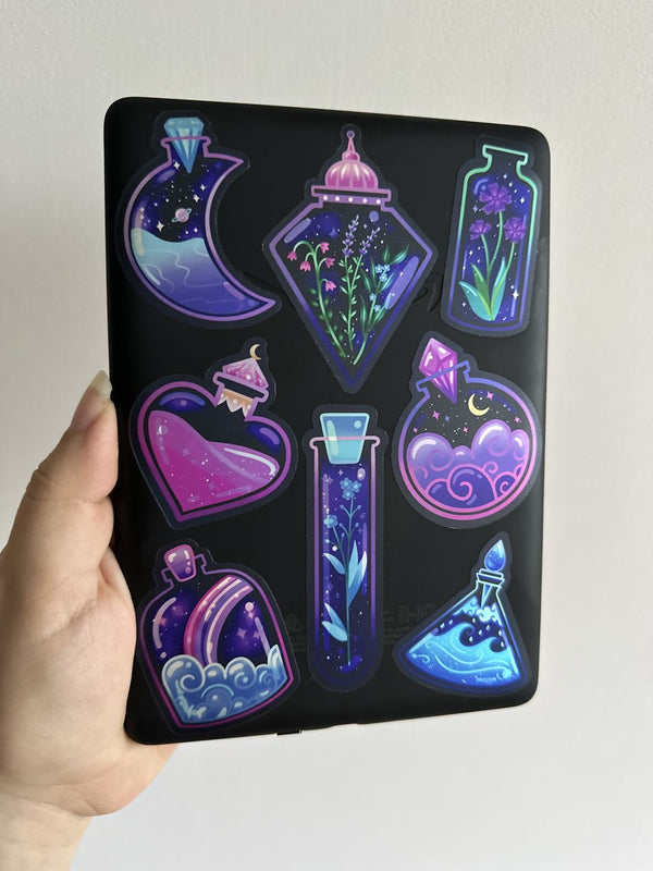 A collection of some The Quirky Cup Collection vinyl stickers in use. A selection of the fantasy potion vinyl stickers in shades of green, blue, pink and purple are stuck to a black notebook.