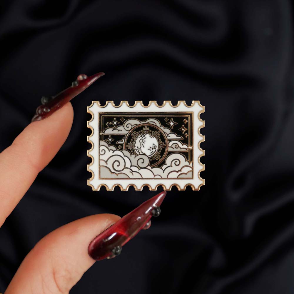 Dark Night- A The Quirky Cup Collective fantasy book inspired enamel pin is held by fingers with red nails above a black background. The black and silver postage stamp enamel pin features moon, cloud and star motifs.