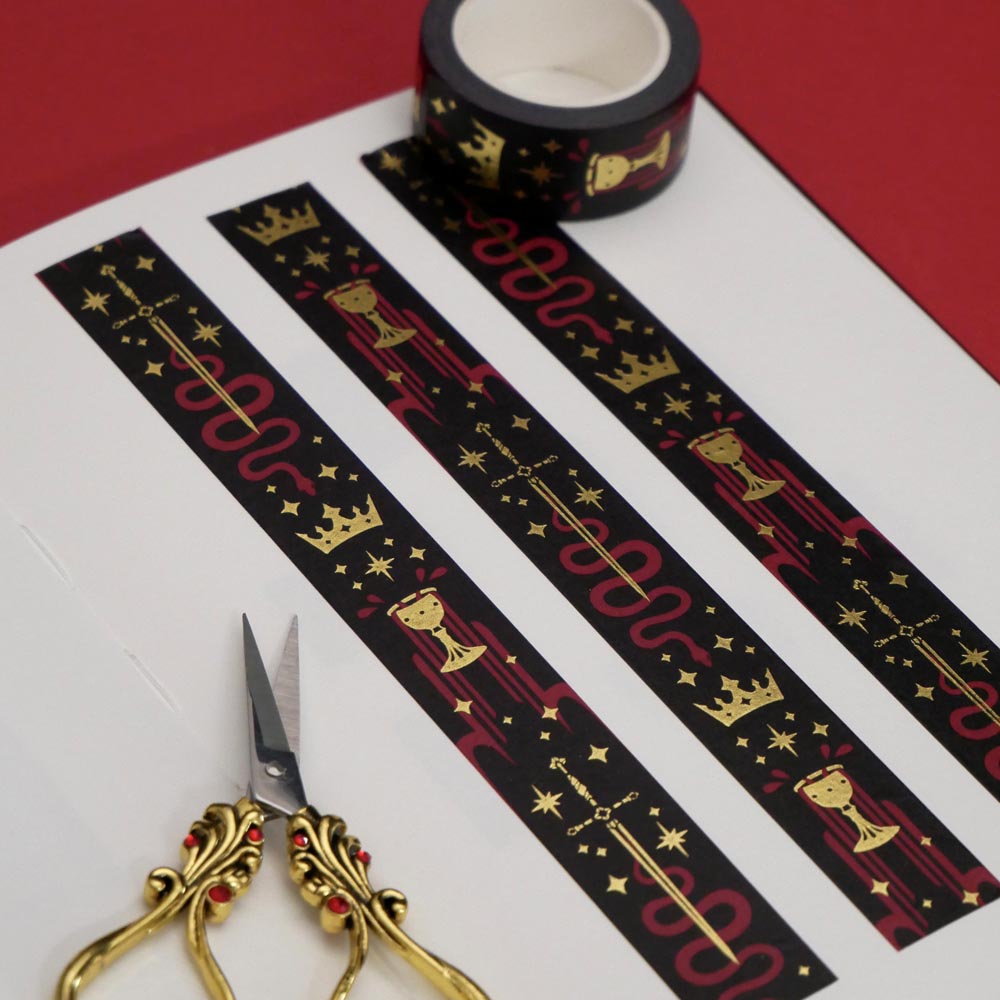 Prophecy Foretold- a fantasy book inspired washi tape by The Quirky Cup Collective- is displayed on journal pages on a red background, small gold and red scissors in the foreground. The bookish washi tape design is black with red and gold detailing- the intricate design showing a crown, red snake, goblet and star icons.