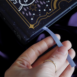 Journal with celestial cover sits on a purple silky background showing the purple foiled edge of the journal and a ribbon bookmark sticking our of the bottom pages being caressed by a hand