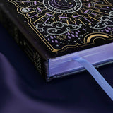 Journal with celestial cover sits on a purple silky background showing the purple foiled edge of the journal and a ribbon bookmark sticking our of the bottom pages