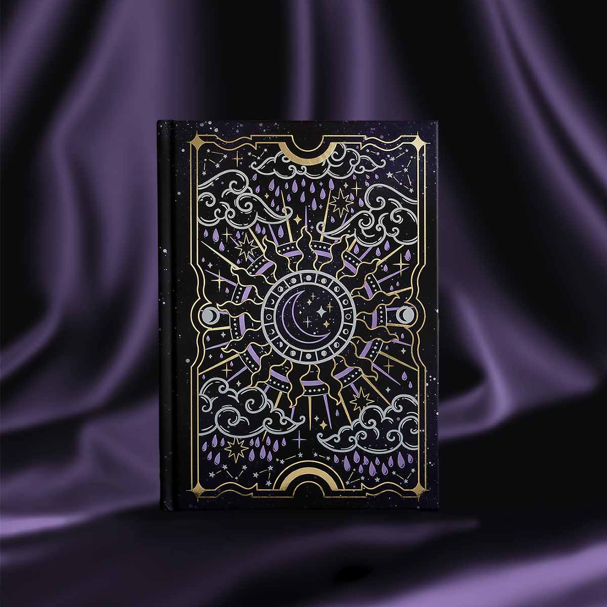 Journal with celestial cover sits on a purple silky background. Journal cover shows a moon nested inside a sun, surrounded by clouds, raindrops and stars. Foiling is purple and gold.