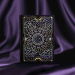 Journal with celestial cover sits on a purple silky background. Journal cover shows a moon nested inside a sun, surrounded by clouds, raindrops and stars. Foiling is purple and gold.