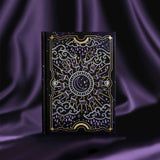 Journal with celestial cover sits on a purple silky background. Journal cover shows a moon nested inside a sun, surrounded by clouds, raindrops and stars. Foiling is purple and gold.