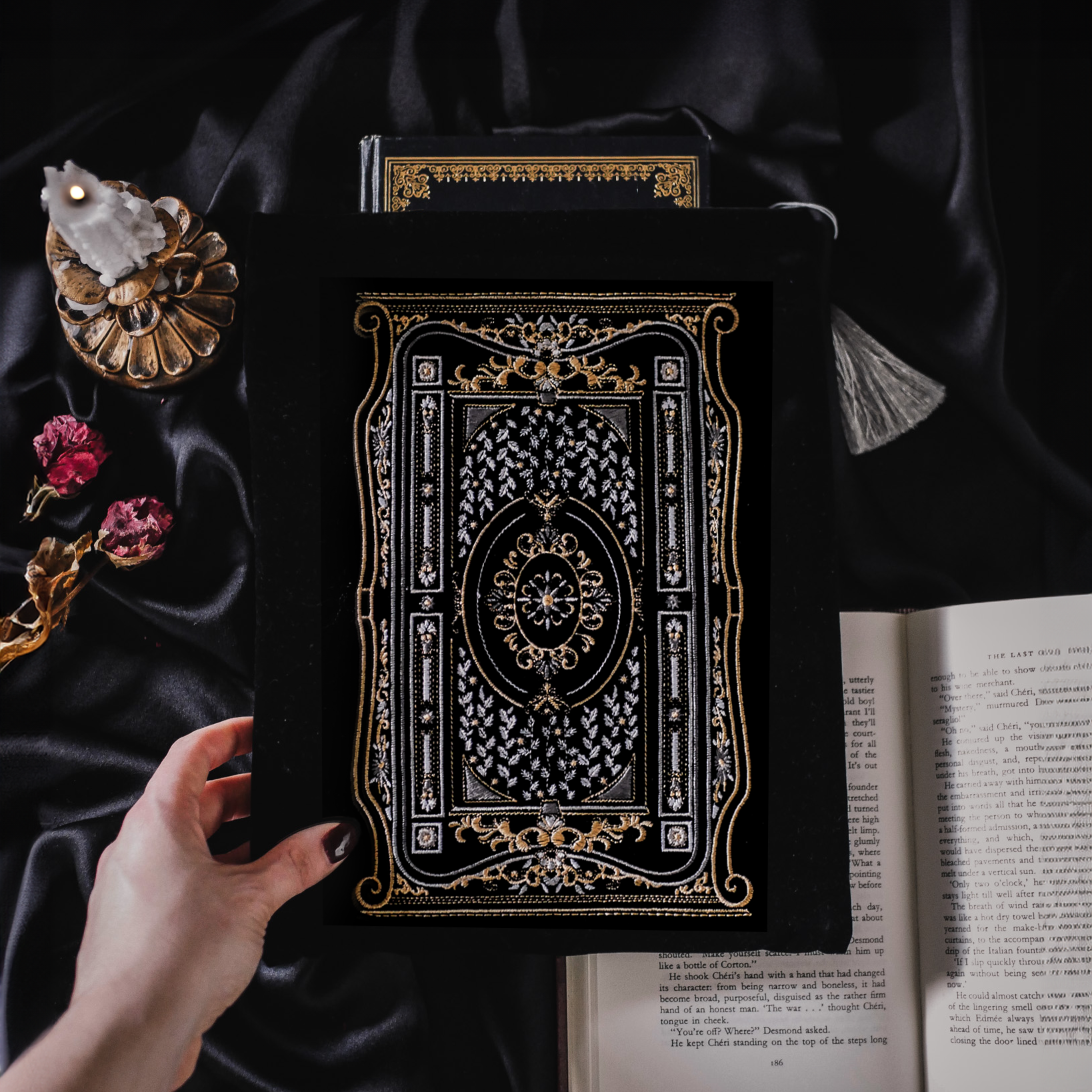 A black book sleeve with an ornate, intricate gold and white design laid on a dark satin fabric. Dried rose petals and an open book are placed next to it, creating a sophisticated, moody atmosphere.