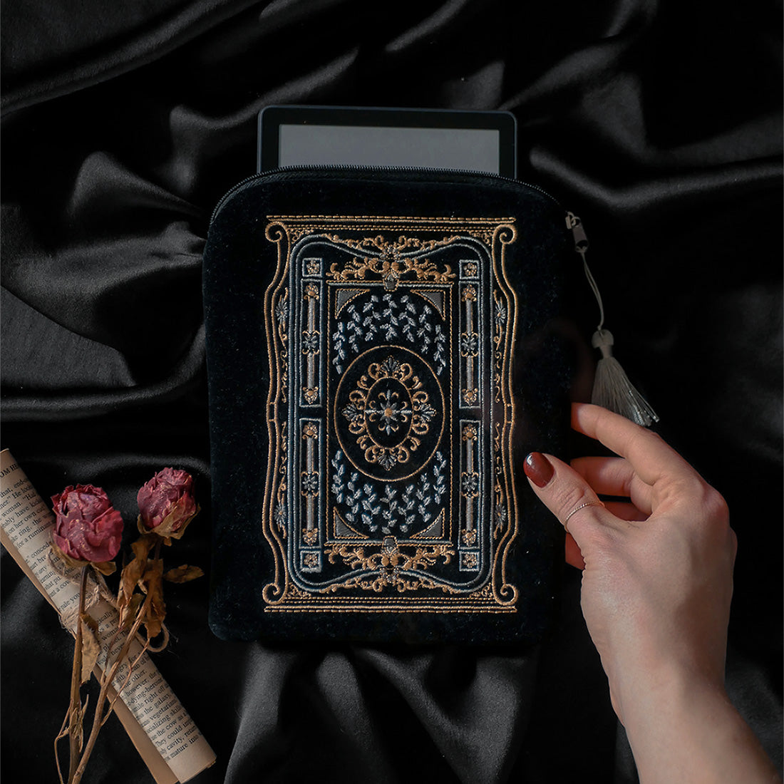 A hand holding a luxurious black velvet book sleeve with an intricate gold and silver embroidered design by The Quirky Cup Collective. The Kindle & E-reader sleeve, featuring a decorative tassel zipper, holds an e-reader, peeking out from the top. The background is a smooth black satin fabric, with dried roses and rolled-up book pages placed beside the Kindle & E-reader sleeve, adding a vintage and elegant aesthetic to the scene.