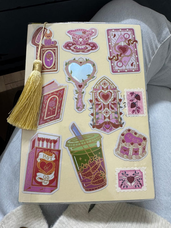 A collection of some The Quirky Cup Collection stickers in use. A selection of book inspired vinyl stickers in shades of pink from the 'Sugar & Spice' collection in sit in a clear kindle case with imagery of strawberry matcha, books, playing cards and stamps.