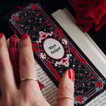 A hand with red nail polish holds a black bookmark with a grid of hearts and roses. The bookmark reads 'Slut for Smut' in an elegant red and white frame. The book it rests on is opened, with a rich red tassel flowing from the bookmark