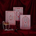 A set of three pink journals with decorative patterns displayed against a red backdrop, accompanied by a glass of wine and a string of pearls.