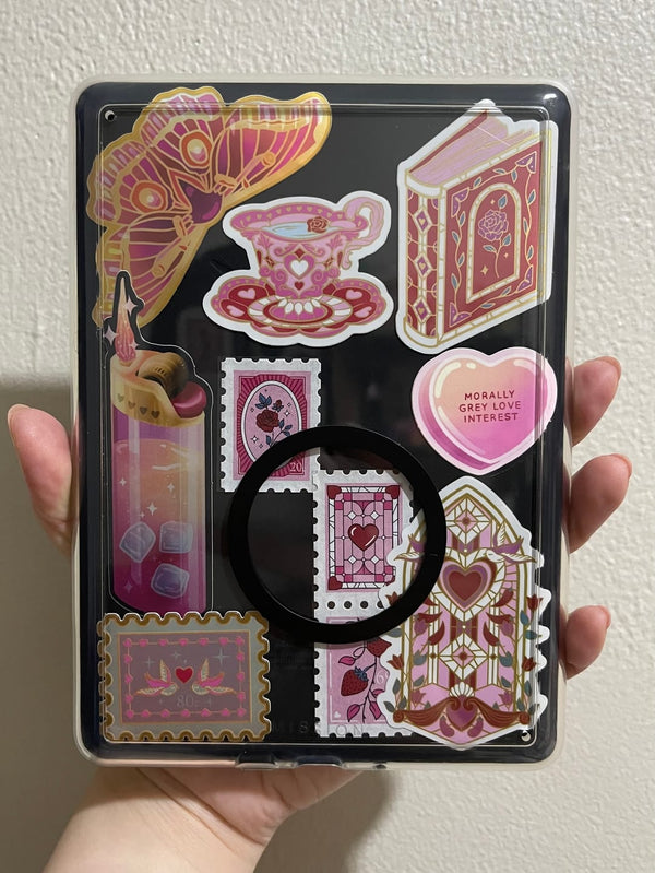 A collection of The Quirky Cup Collection bookish stickers in use. A selection of booktok inspired vinyl stickers in pink and gold tones sit on a black e-reader underneath a clear case.
