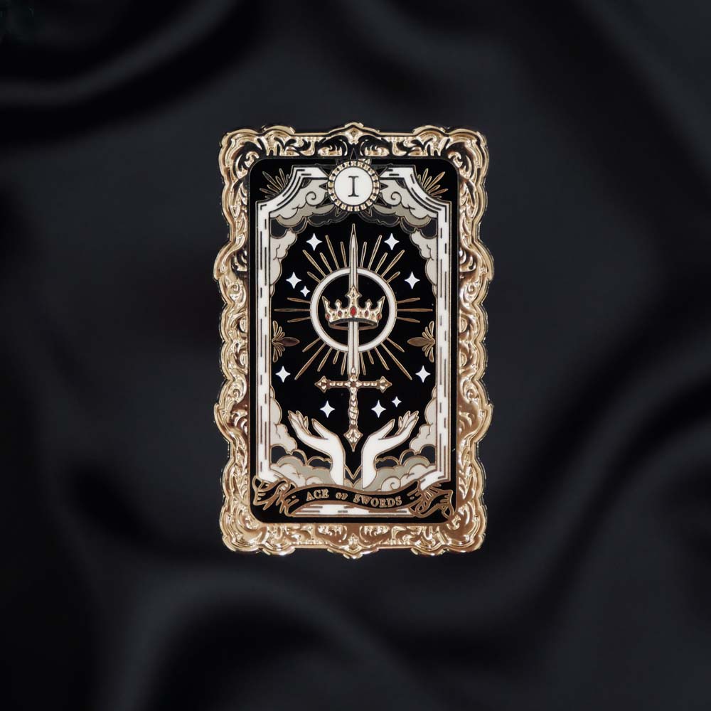 The fantasy book inspired Ace of Swords enamel pin by The Quirky Cup Collective sits on a black background. The gold pin features black and red detailing and shows a tarot card style illustration with a crown and sword motif amongst clouds.
