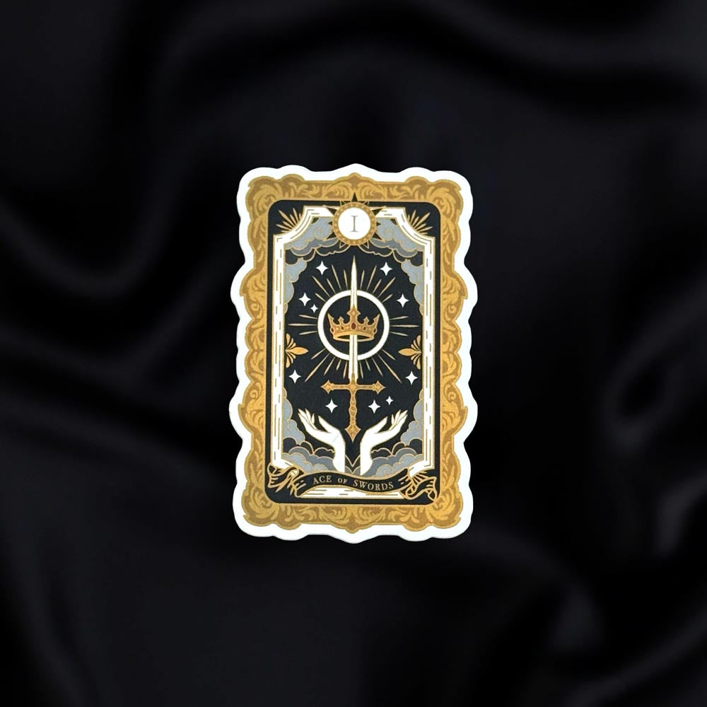 The Ace of Swords- a fantasy vinyl sticker by The Quirky Cup Collective as part of the 'Serpents and Swords' collection- sits on a black fabric background. The book inspired sticker shows a tarot-card inspired design of hands reaching towards a crown and sword in shades of black, cold and silver.