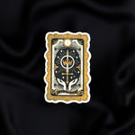 The Ace of Swords- a fantasy vinyl sticker by The Quirky Cup Collective as part of the 'Serpents and Swords' collection- sits on a black fabric background. The book inspired sticker shows a tarot-card inspired design of hands reaching towards a crown and sword in shades of black, cold and silver.