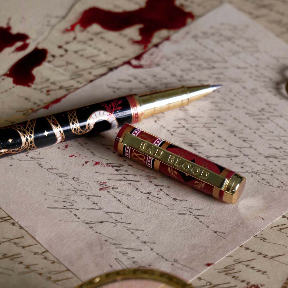 The Quirky Cup Collective Bad Blood pen features an intricate snake motif in red, gold and black and an engraved 'Bad Blood' text on the clip. The pen sits on a background of handwritten calligraphy notes and spilt red wax.
