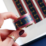 A close up of the Blood Spilled washi tape by The Quirky Cup Collective. The roll of fantasy inspired washi tape is held by a hand in front of a journal showcasing the fantasy inspired stationery. The fantasy book inspired washi tape design is pink, blue with gold detailing with a goblet motif.