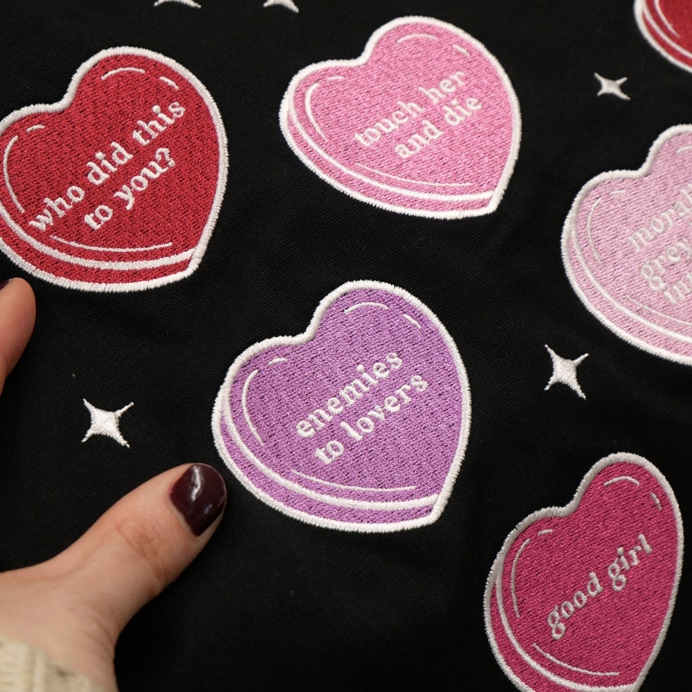 a close up of a black canvas tote bag covered in candy hearts and stars. The stars are in shades of purple, pink and red and refer to common romance fantasy book tropes