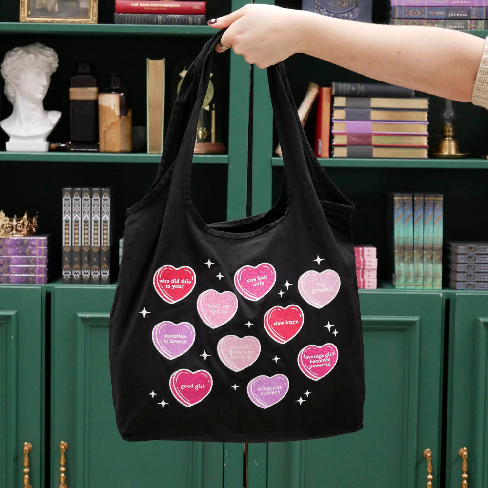 a hand holds a black canvas tote bag covered in candy hearts and stars. The stars are in shades of purple, pink and red and refer to common romance fantasy book tropes