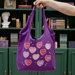 woman holds purple canvas tote bag covered in candy hearts and stars. The stars are in shades of purple, pink and red and refer to common romance fantasy book tropes
