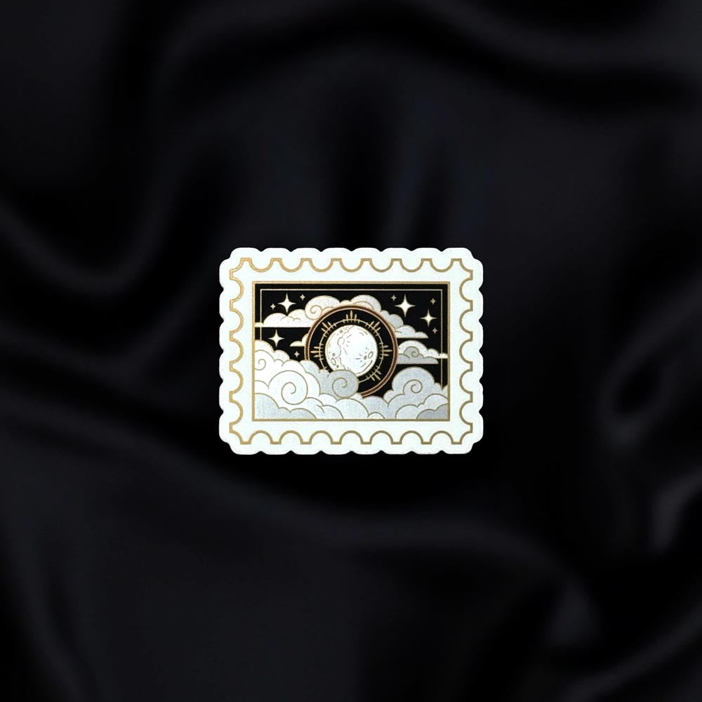Dark Night Stamp- a fantasy vinyl sticker by The Quirky Cup Collective as part of the 'Serpents and Swords' collection- sits on a black fabric background. The fantasy book sticker in the shape of a postage stamp shows the moon in front of a silver and black cloudy sky filled with stars.