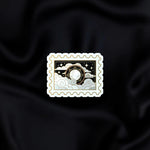 Dark Night Stamp- a fantasy vinyl sticker by The Quirky Cup Collective as part of the 'Serpents and Swords' collection- sits on a black fabric background. The fantasy book sticker in the shape of a postage stamp shows the moon in front of a silver and black cloudy sky filled with stars.