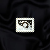 Dark Night Stamp- a fantasy vinyl sticker by The Quirky Cup Collective as part of the 'Serpents and Swords' collection- sits on a black fabric background. The fantasy book sticker in the shape of a postage stamp shows the moon in front of a silver and black cloudy sky filled with stars.