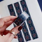 A close up of the Dragon Bornwashi tape by  The Quirky Cup Collective. The roll of fantasy inspired washi tape is held by a hand in front of a journal showcasing the fantasy inspired stationery. The bookish washi tape design is blue, pink with gold detailing- the intricate design showing a serpent eye.