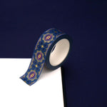 A close up of the Dragon Bornwashi tape by The Quirky Cup Collective. The roll of fantasy inspired washi tape is on the corner of a white journal on a blue background. The bookish washi tape design is blue, pink with gold detailing- the intricate design showing a serpent eye.