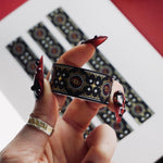 A close up of the Dragon Born washi tape by The Quirky Cup Collective. The roll of fantasy inspired washi tape is held in a hand with intricate red nails over a journal on a red table. The bookish washi tape design is red and black with gold detailing- the intricate design showing a serpent eye.