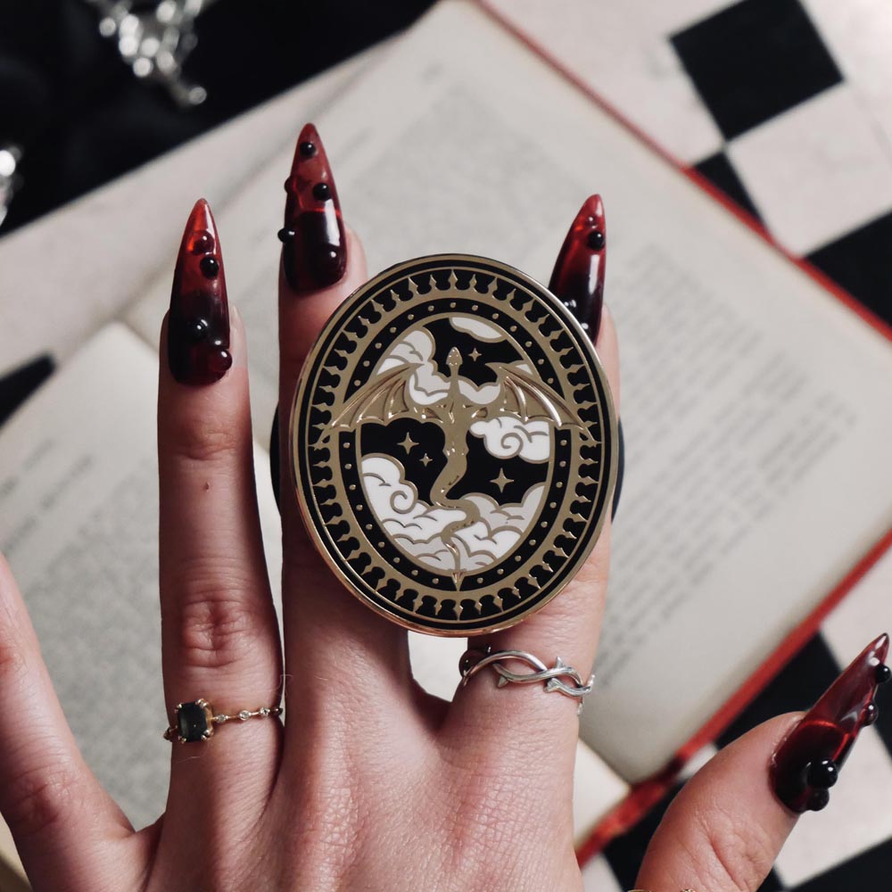 The Dragon Lore clingie by The Quirky Cup Collective is held by red nailed fingers above a red book and gaming table. The gold kindle grip features black,white and gold detailing and shows a dragon flying over clouds with a decorative border in a oval shape.