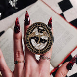 The Dragon Lore clingie by The Quirky Cup Collective is held by red nailed fingers above a red book and gaming table. The gold kindle grip features black,white and gold detailing and shows a dragon flying over clouds with a decorative border in a oval shape.