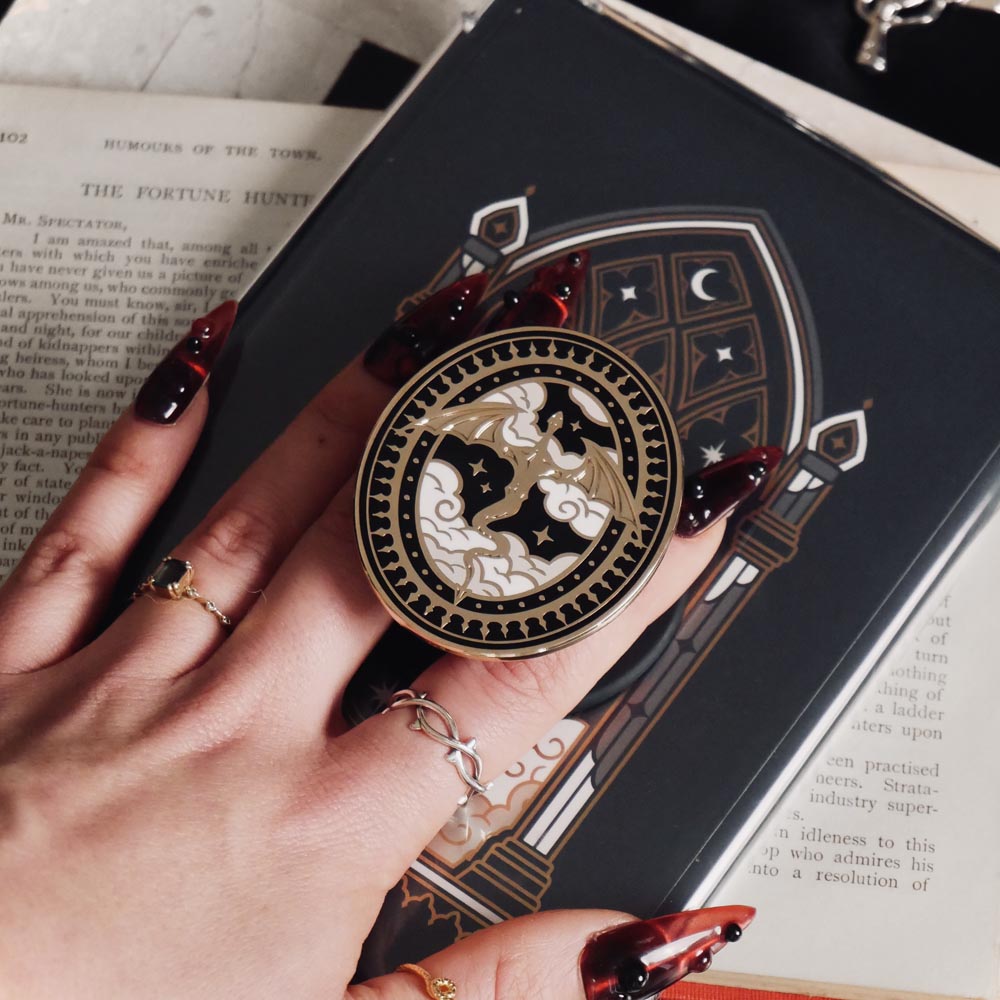 The Dragon Lore clingie by The Quirky Cup Collective is held by red nailed fingers as it is attached to a matching kindle sticker insert. The gold kindle grip features black,white and gold detailing and shows a dragon flying over clouds with a decorative border in a oval shape.