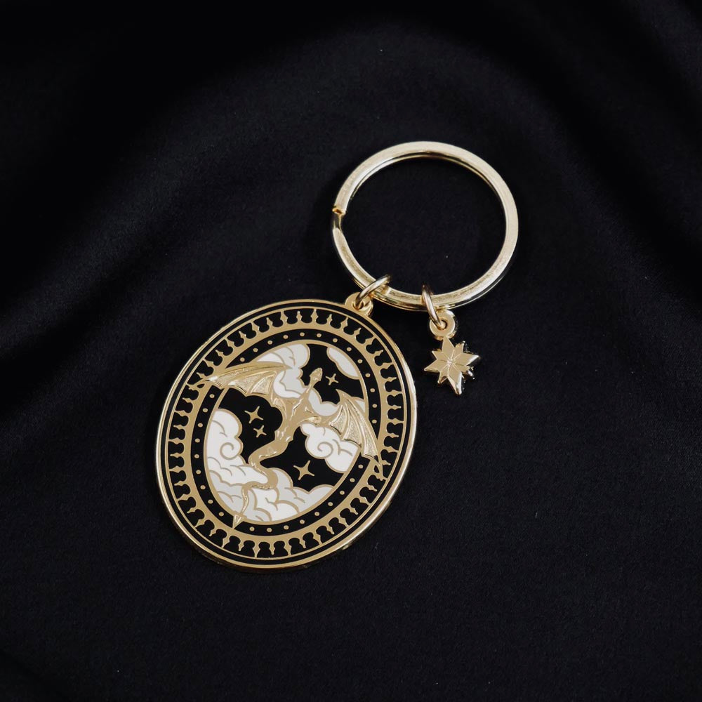 The Dragon Lore fantasy book inspired keyring by The Quirky Cup Collective sits on a black background. The gold keyring features black,white and gold detailing and shows a dragon flying over clouds with a decorative border, a small star charm also dangling from the keychain.