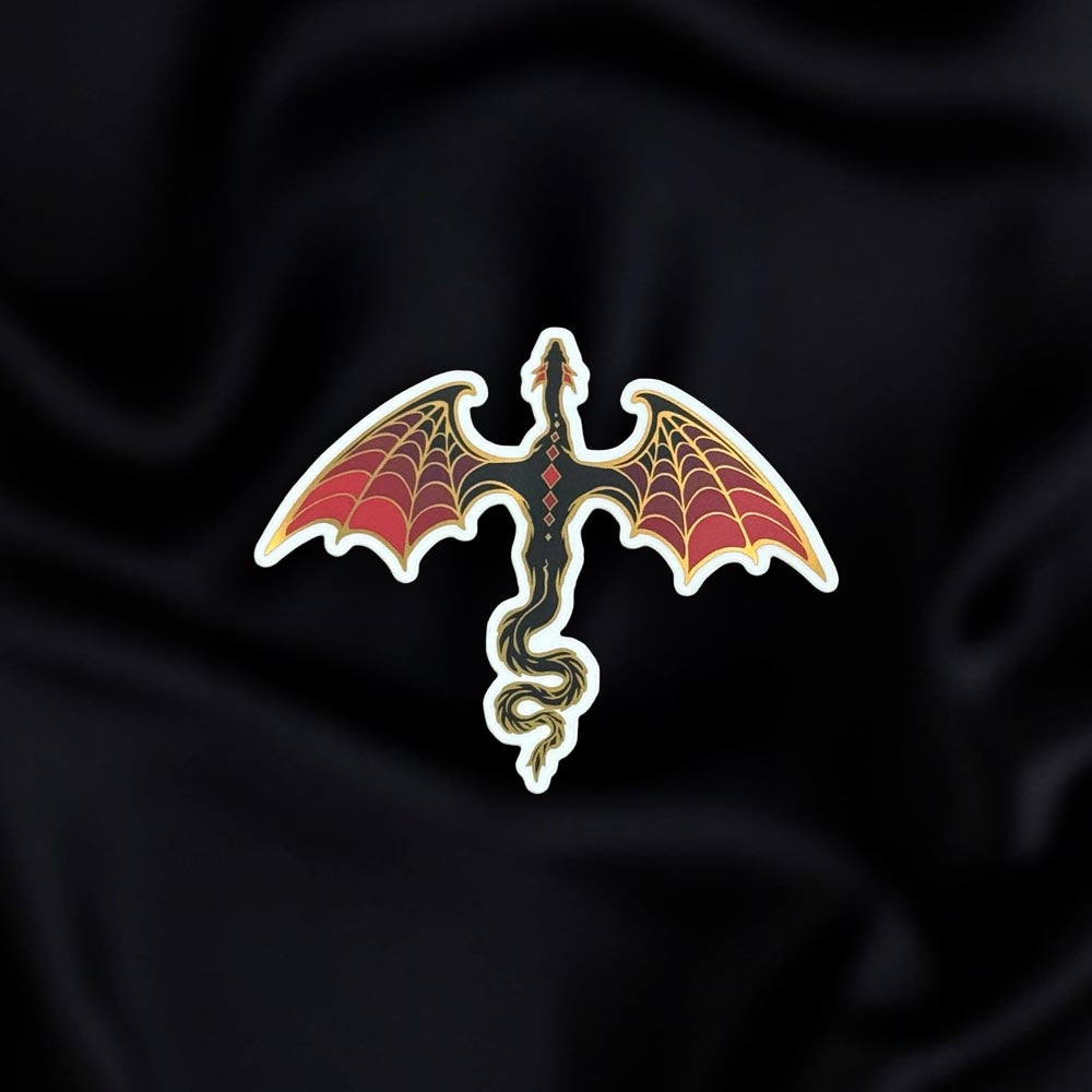 Dragon Rider- a fantasy vinyl sticker by The Quirky Cup Collective as part of the 'Serpents and Swords' collection- sits on a black fabric background. The fantasy book sticker shows a black and red dragon with gold detailing flying, as viewed from above.