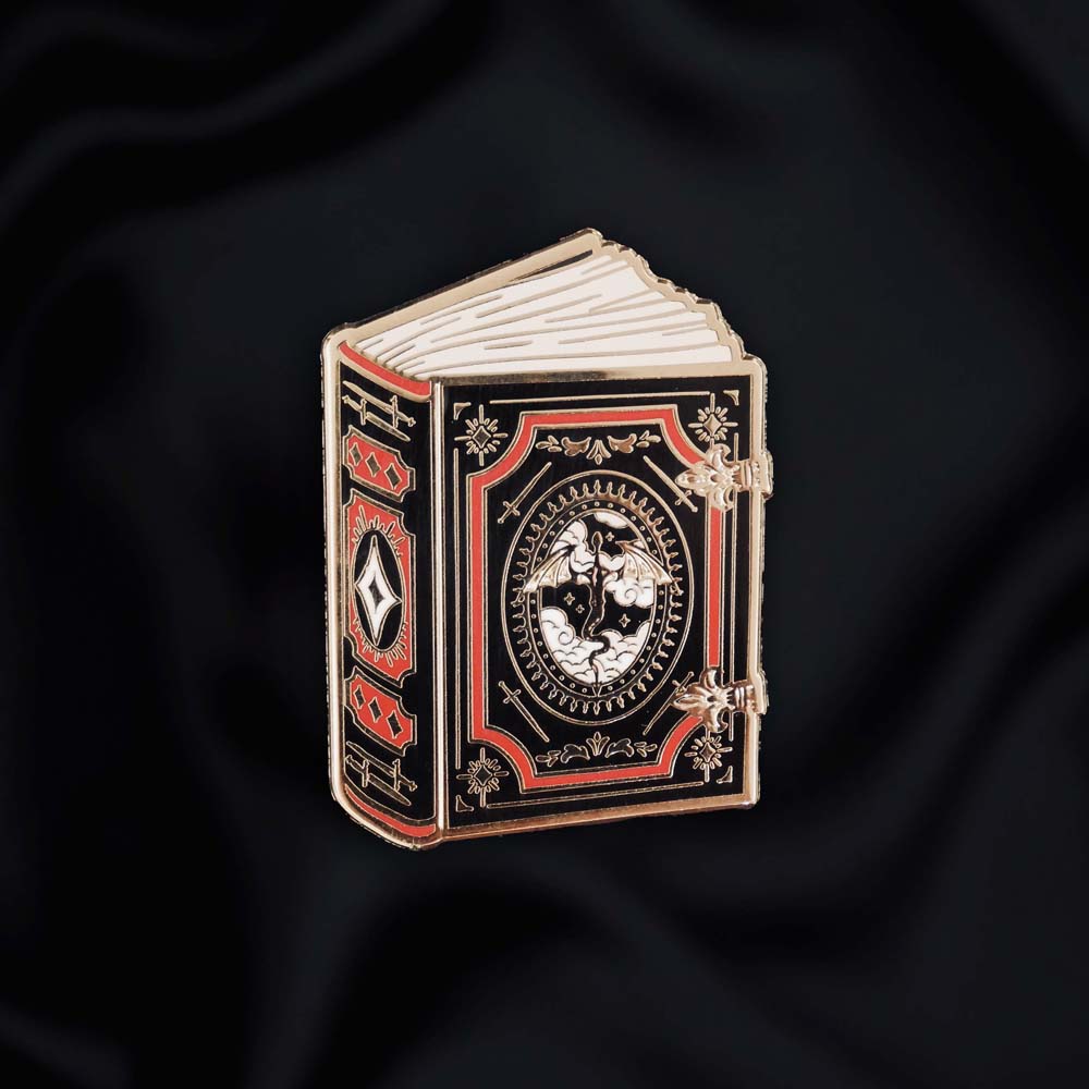 black red and gold book with dragon enamel pin held by a hand against a dark background