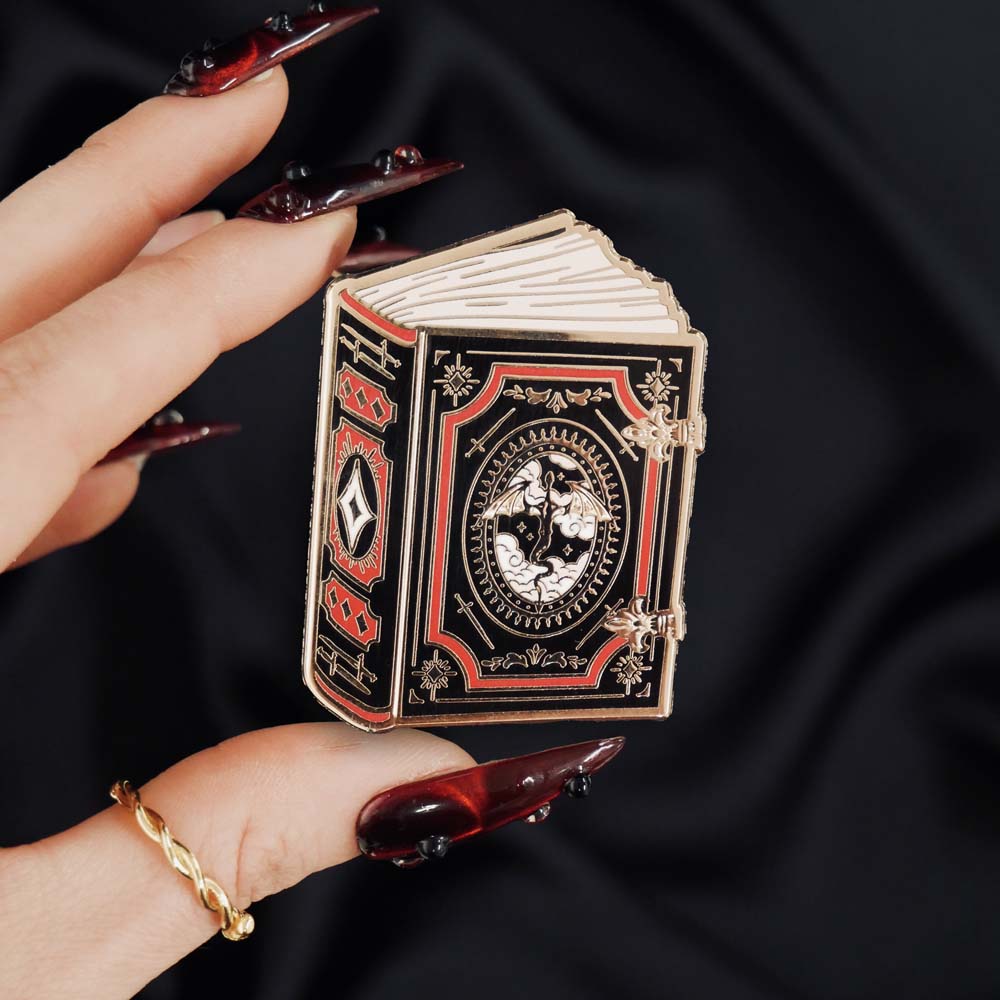 black red and gold book with dragon enamel pin held by a hand against a dark background
