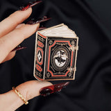 black red and gold book with dragon enamel pin held by a hand against a dark background