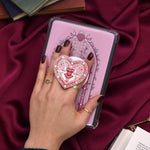 A hand holds the pink Everything Nice clingie on a kindle with a kindle sticker insert over a red fabric background. The kindle sticker insert design centers on a light pink stained glass arch design in shades of pink, red and green, the clingie featuring a heart and birds in a heart shaped design.