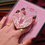 Heart-shaped E-reader and phone grip held by a hand with dark nail polish against a pink background