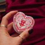 The Everything Nice clingie is held in a hand in front of a red fabric background. The enamel kindle grip features heart and birds surrounded by flowers in a heart shaped design.