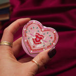 Heart-shaped pink phone & kindle grip with decorative design held by a hand against a red fabric background