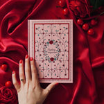 The Reading Companion - A pink journal laying across a draped red silk backdrop surrounded by roses and cherries. Pink Journal is inpspired by stained glass and includes roses and vines, centred with 'Reading Companion' text at the centre