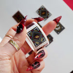 Serpents and Swords- a fantasy book inspired stamp washi tape by The Quirky Cup Collective- is held by a red nailed hand over a journal. The bookish washi tape design features a postage stamp diecut and black, red, gold and white designs with motifs like a crown, snake, goblet, sword and stars.