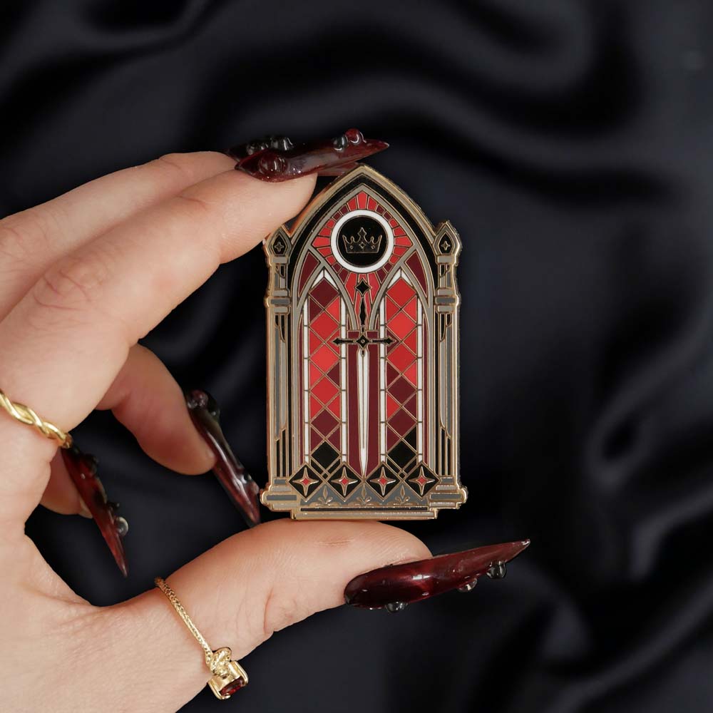 black red and gold window with sword and crown enamel pin held by a hand against a dark background