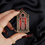 black red and gold window with sword and crown enamel pin held by a hand against a dark background