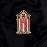 black red and gold window with sword and crown enamel pin held by a hand against a dark background