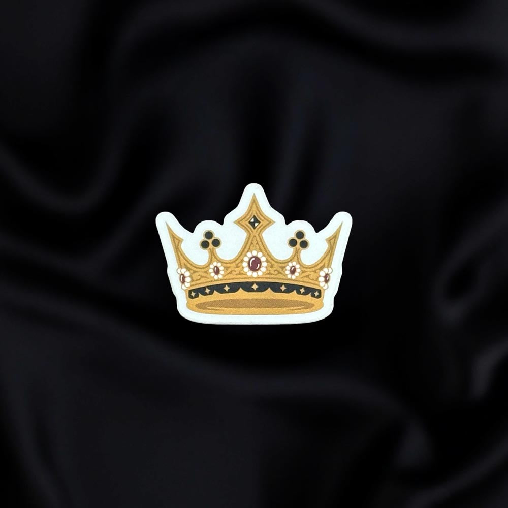 Gilded Heir- a fantasy vinyl sticker by The Quirky Cup Collective as part of the 'Serpents and Swords' collection- sits on a black fabric background. The fantasy book sticker shows a gold grown with white, red and black detailing.