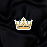 Gilded Heir- a fantasy vinyl sticker by The Quirky Cup Collective as part of the 'Serpents and Swords' collection- sits on a black fabric background. The fantasy book sticker shows a gold grown with white, red and black detailing.