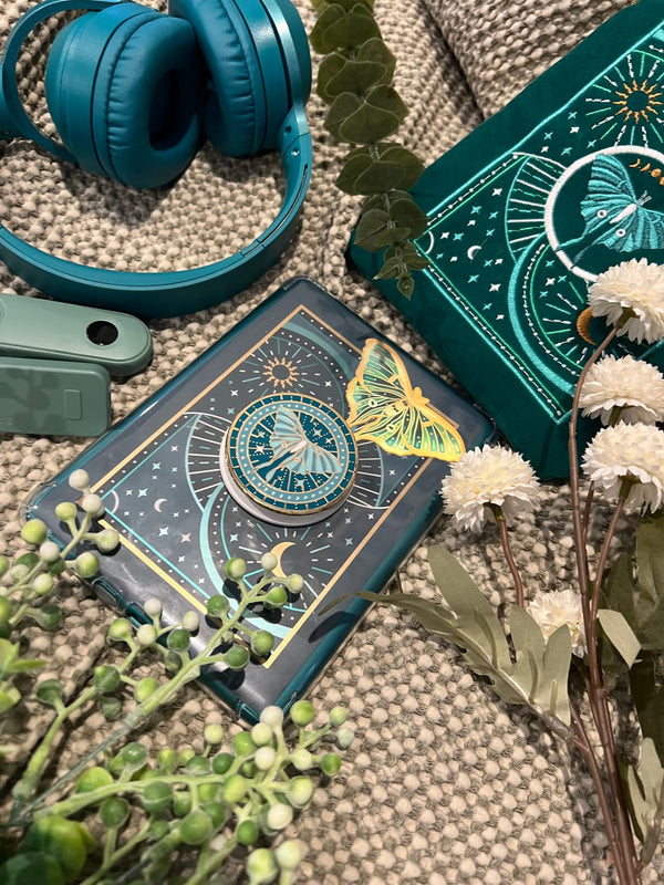 A collection The Quirky Cup Collection book inspired products together. A clear kindle case contains the Luna moth kindle sticker insert alongside a gold luna moth sticker. A Luna moth clingie kindle grip sits on the outside with a embroidered luna moth ereader soft case also in the scene.