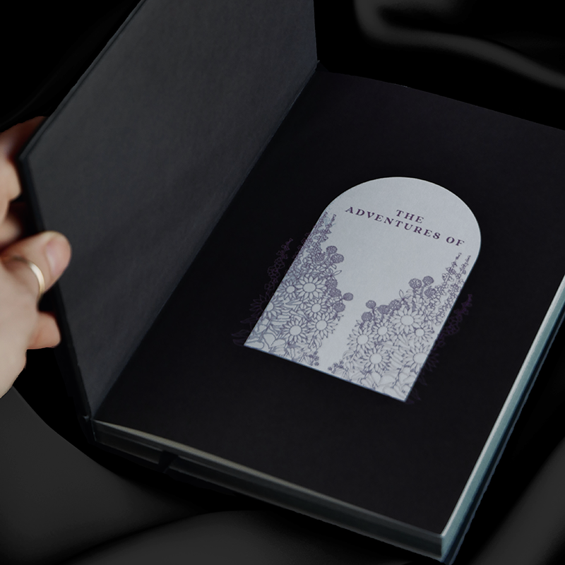 black journal sits open on background of black draped silk. On the front end page it says 'the adventures of' to show where you can write your name