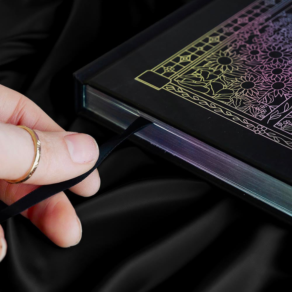 decorative black journal spine with holographic foiling sits on a background of black draped silk. Hand is holding a bookmark ribbon coming out of journal
